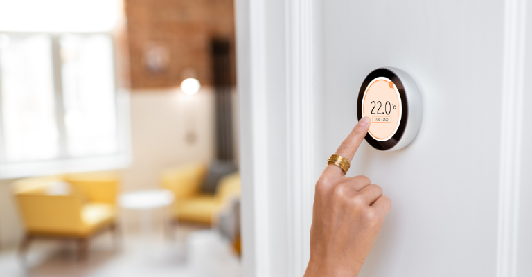 A person adjusts a smart thermostat on a white wall, setting the temperature to 22 degrees Celsius; a lounge with yellow chairs is visible in the background.