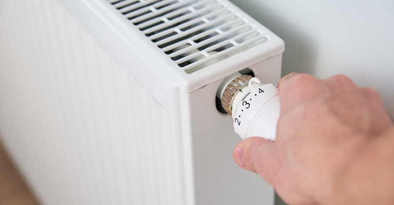 A hand adjusts the temperature dial on a white radiator mounted against a light-coloured wall.