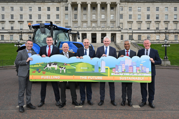 Seven men in suits stand outside a government building, holding a colourful banner that reads "Fuelling the Future Sustainably" with illustrations of farming and renewable energy.